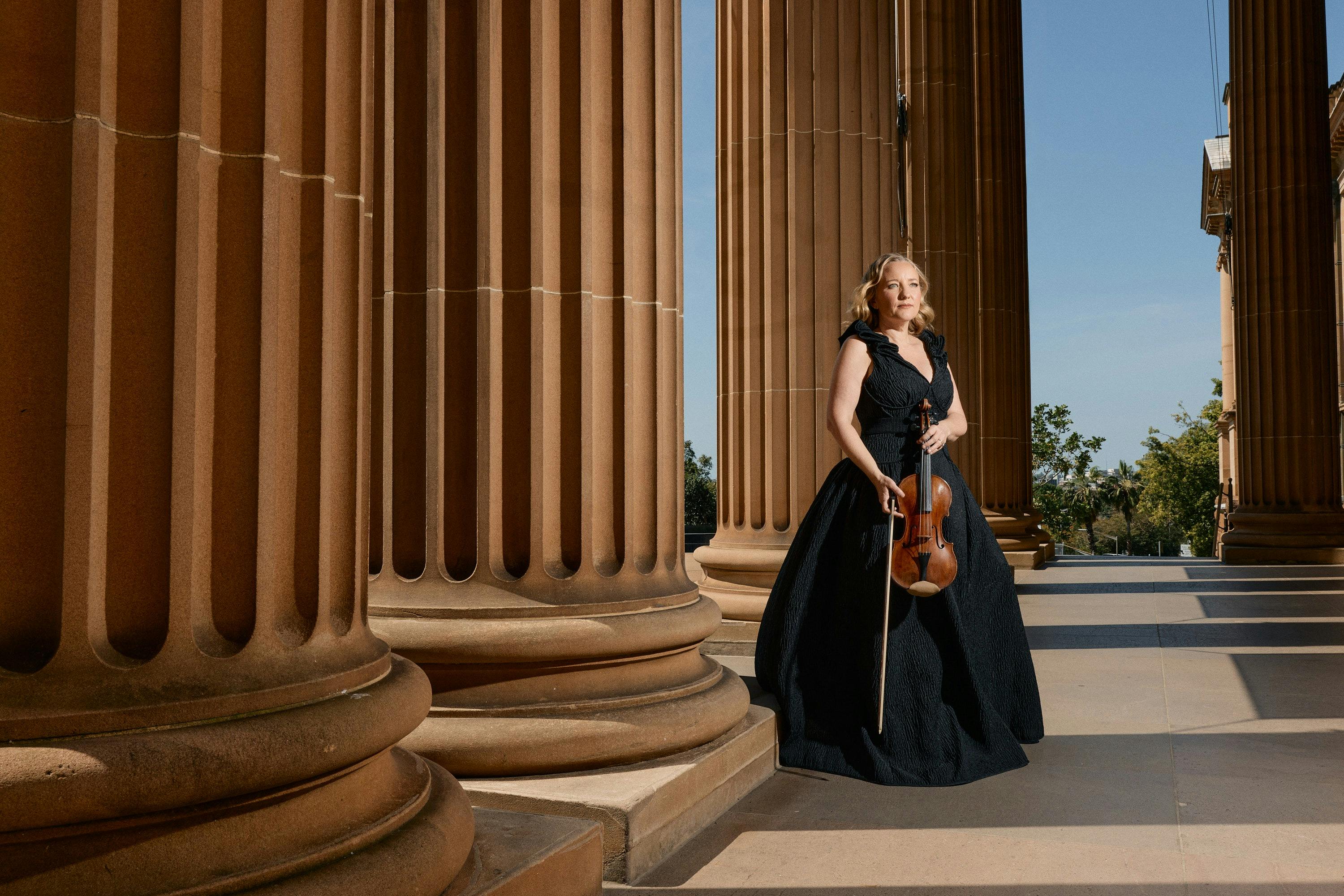 A woman in formalwear holding a violin posing beside sandstone columns