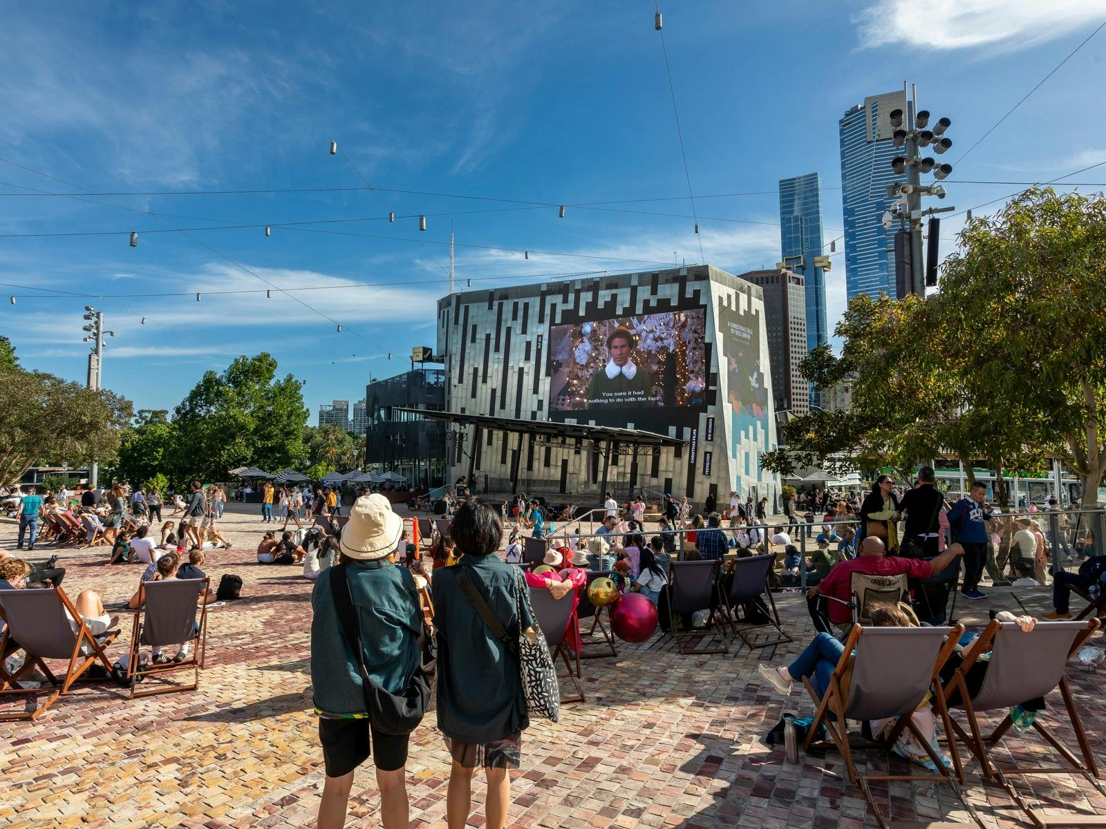 Crowds of people sit on deckchairs watching the film 'Elf' on the big screen at Fed Square