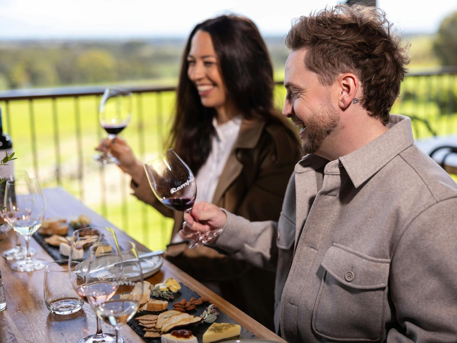 A man and women smiling at their glasses of wine and eating a native tasting platter