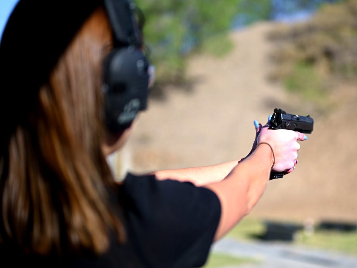 Woman at an outdoor shooting range aiming a handgun wearing protective earmuffs