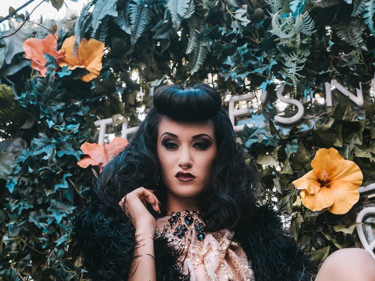 a headshot of a woman with black hair and a fur coat, in front of greenery