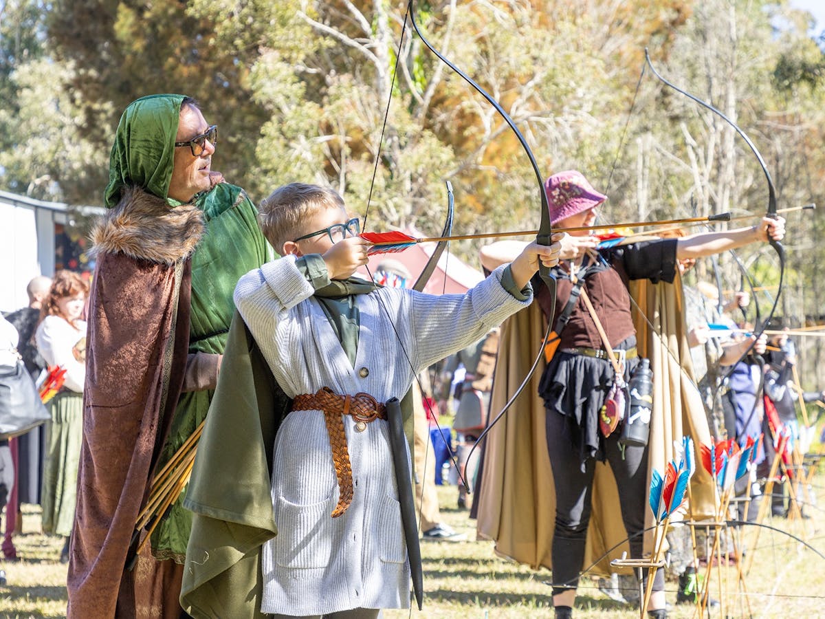Visitors dressed in medieval costumes are drawing a bow and arrow ready to fire.