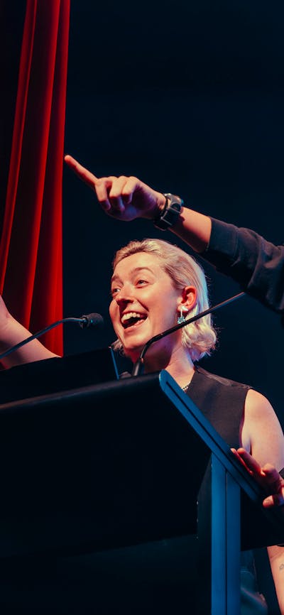 Hosts Harriet Gillies and Xanthe Dobbie on-stage behind a lectern, joyously pointing at audience.