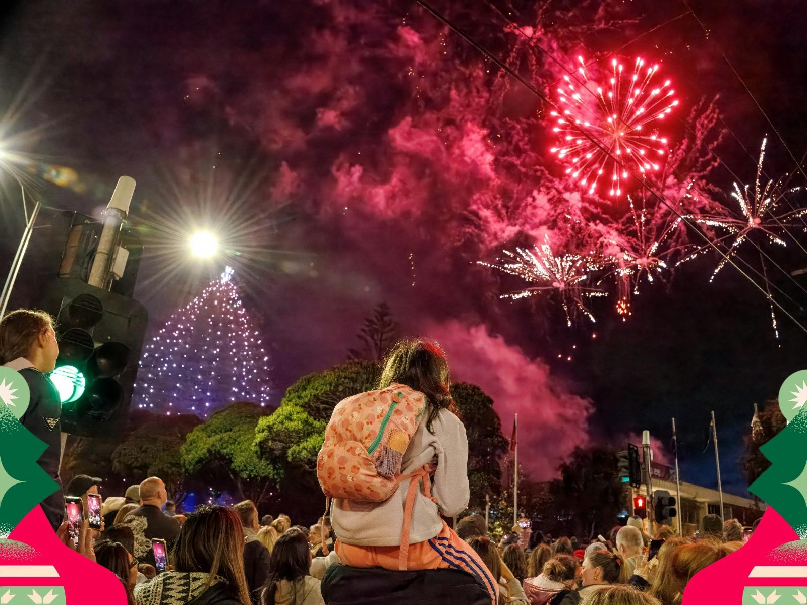 Crowd watches fireworks above a lit Christmas tree, with a child sitting on someone's shoulders.
