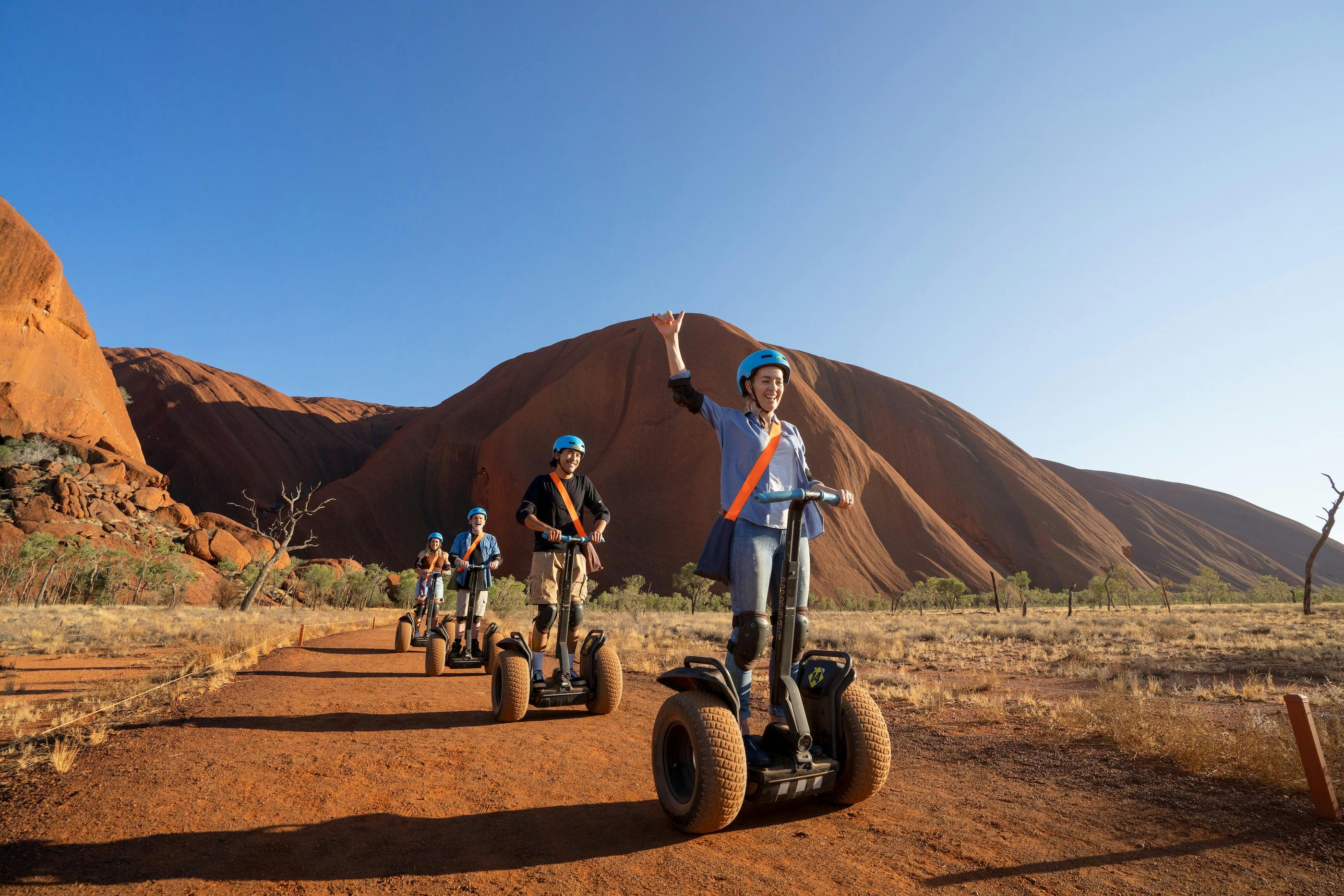 A lady Segways on a path with Uluru a vivid red behind her contrasting against an endless blue sky.