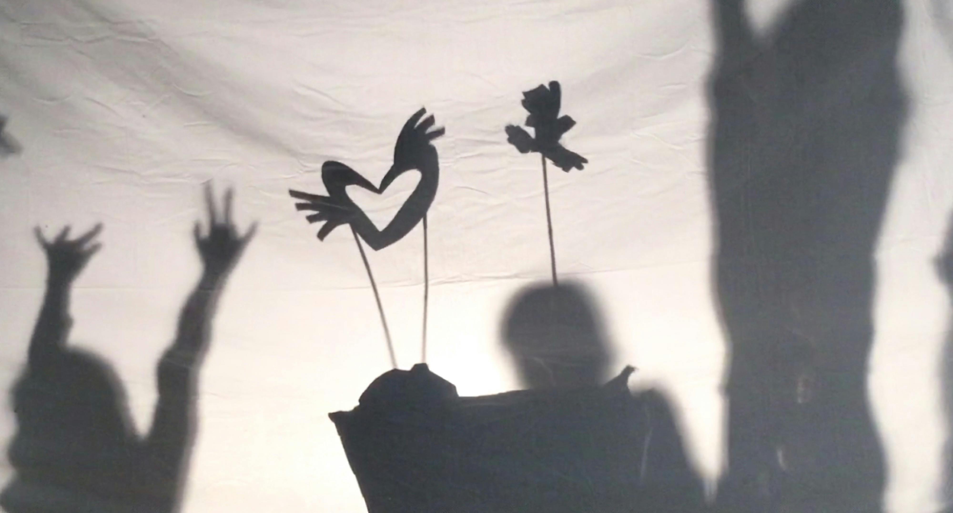 Shadow puppets move across a screen as the outlines of children dancing are visible on either side