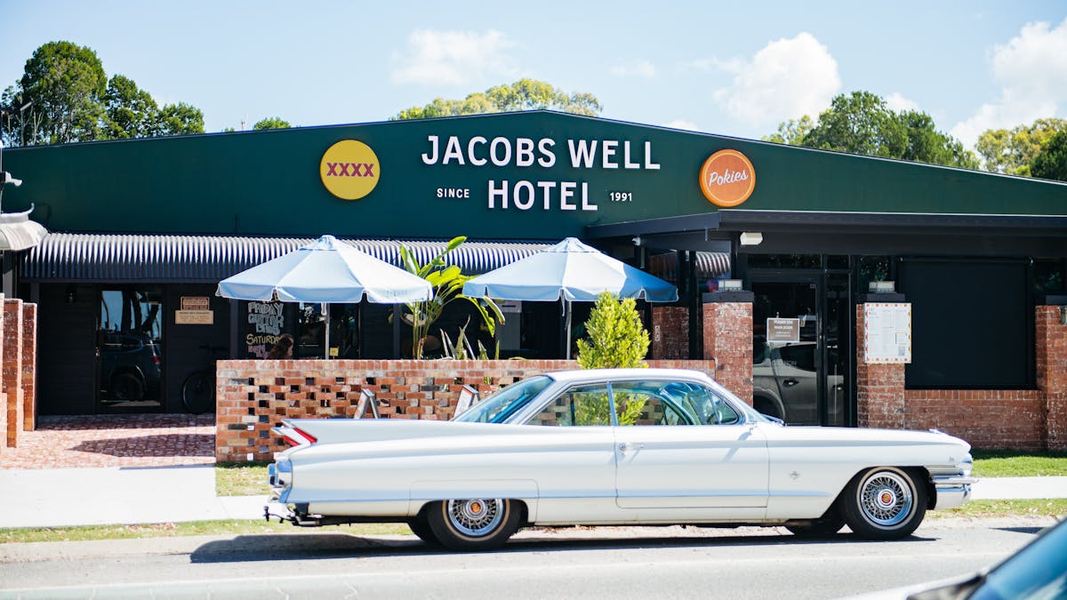 A vintage white Cadillac parked in front of the dark green Jacobs Well Hotel building on a sunny day