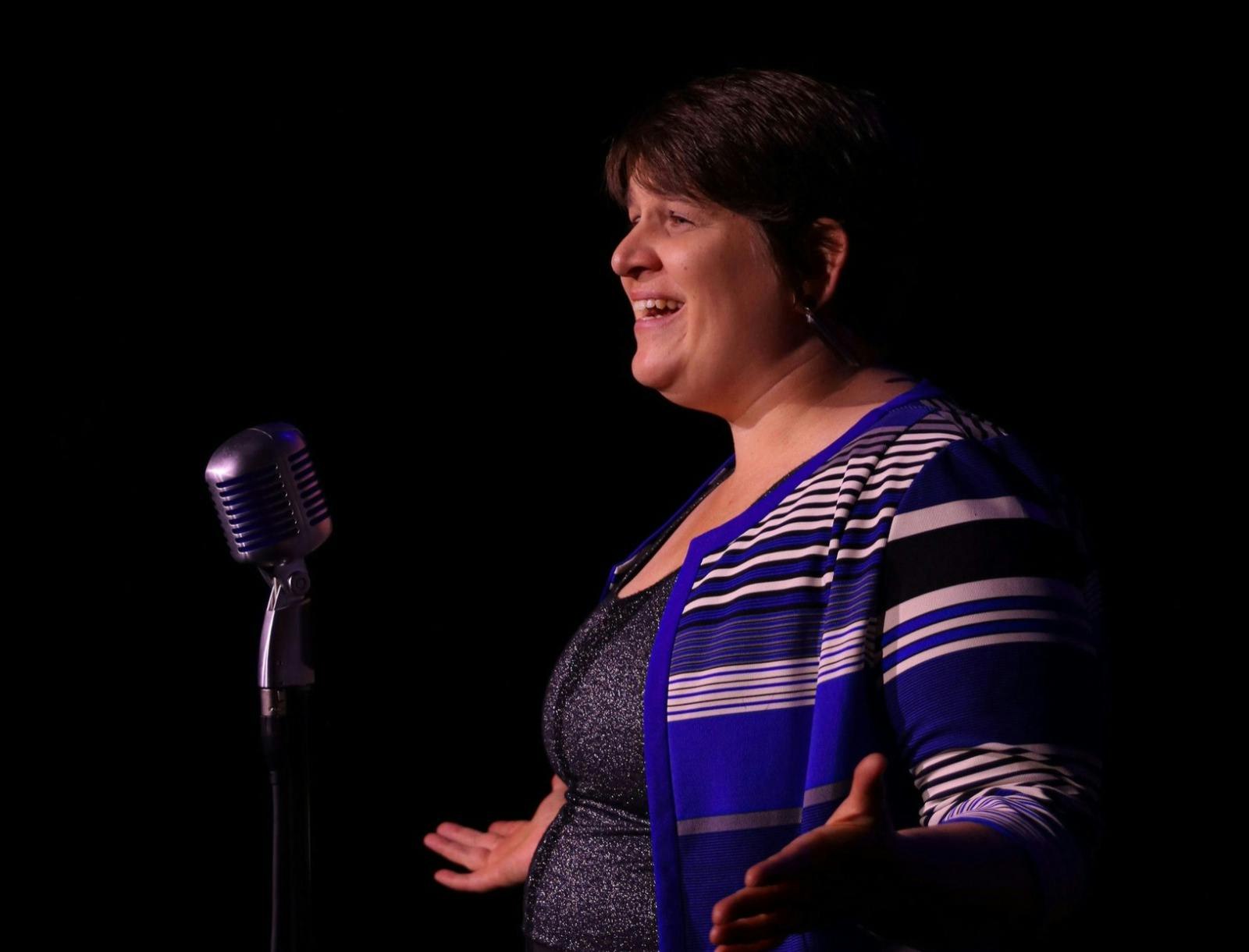 Megan J Riedl smiles while speaking into a vintage-style microphone on stage. She wears a blue, blac