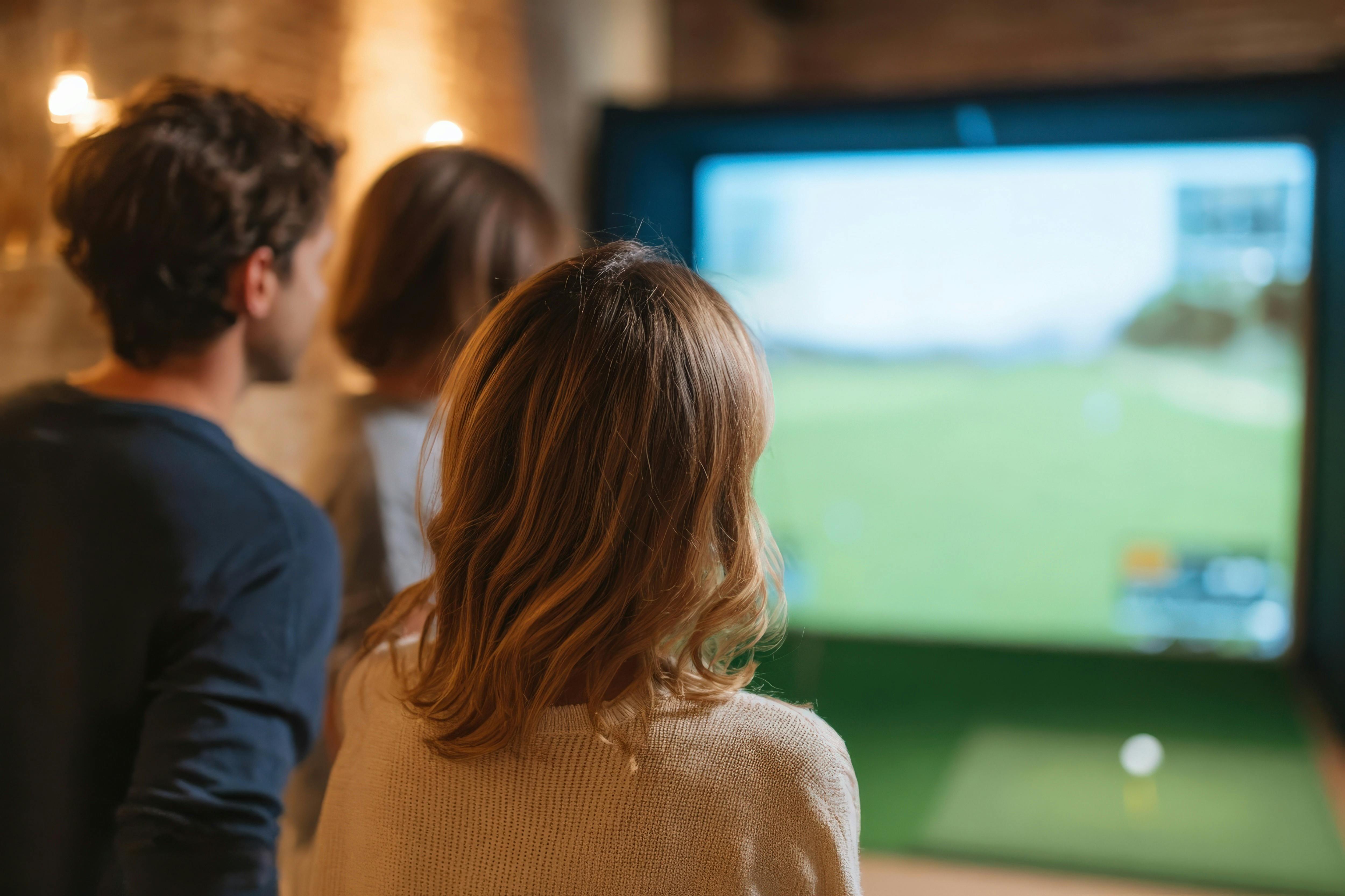 Three people watching a golf simulation on a large indoor screen from behind.