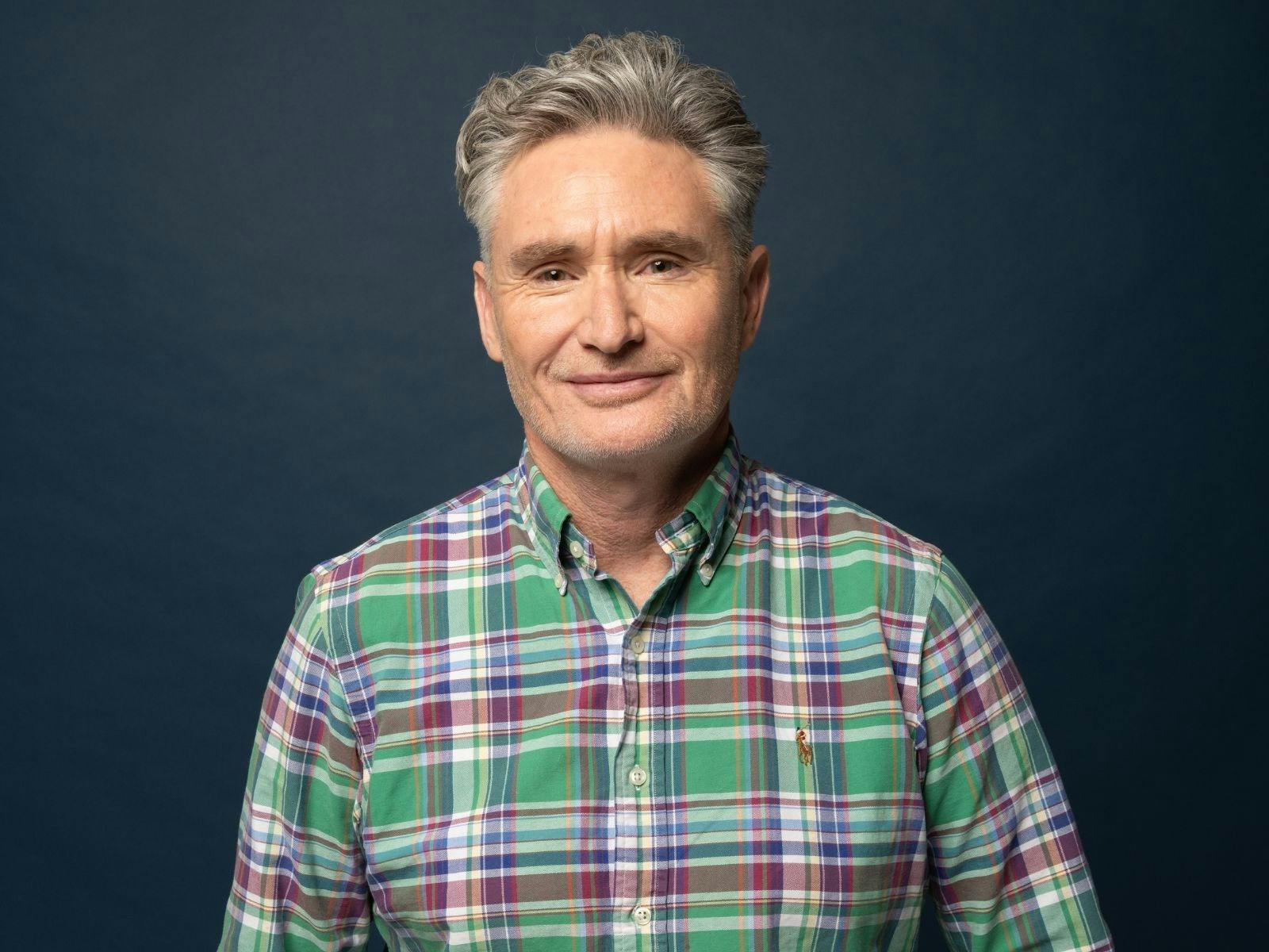 Dave Hughes in a plaid shirt smirking in front of a dark blue wall