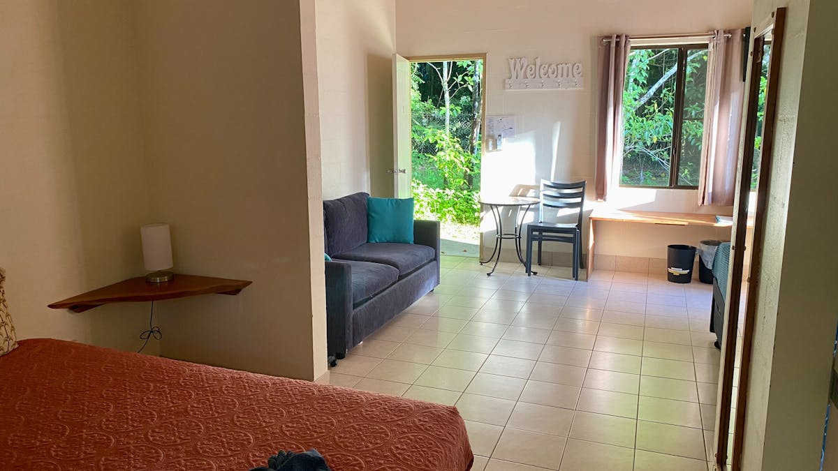 Motel style room showing edge of queen bed, sofa, tiled floor and green rainforest out back door