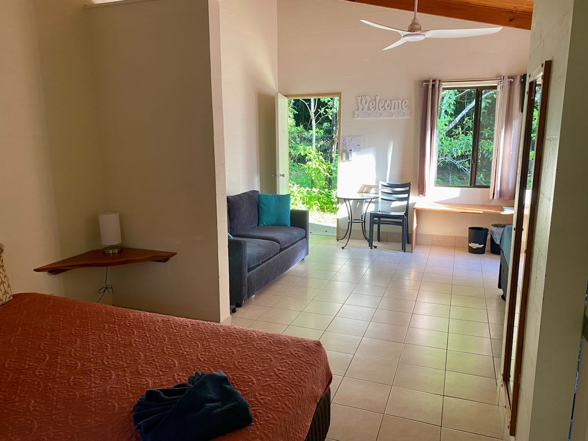 Motel style room showing edge of queen bed, sofa, tiled floor and green rainforest out back door