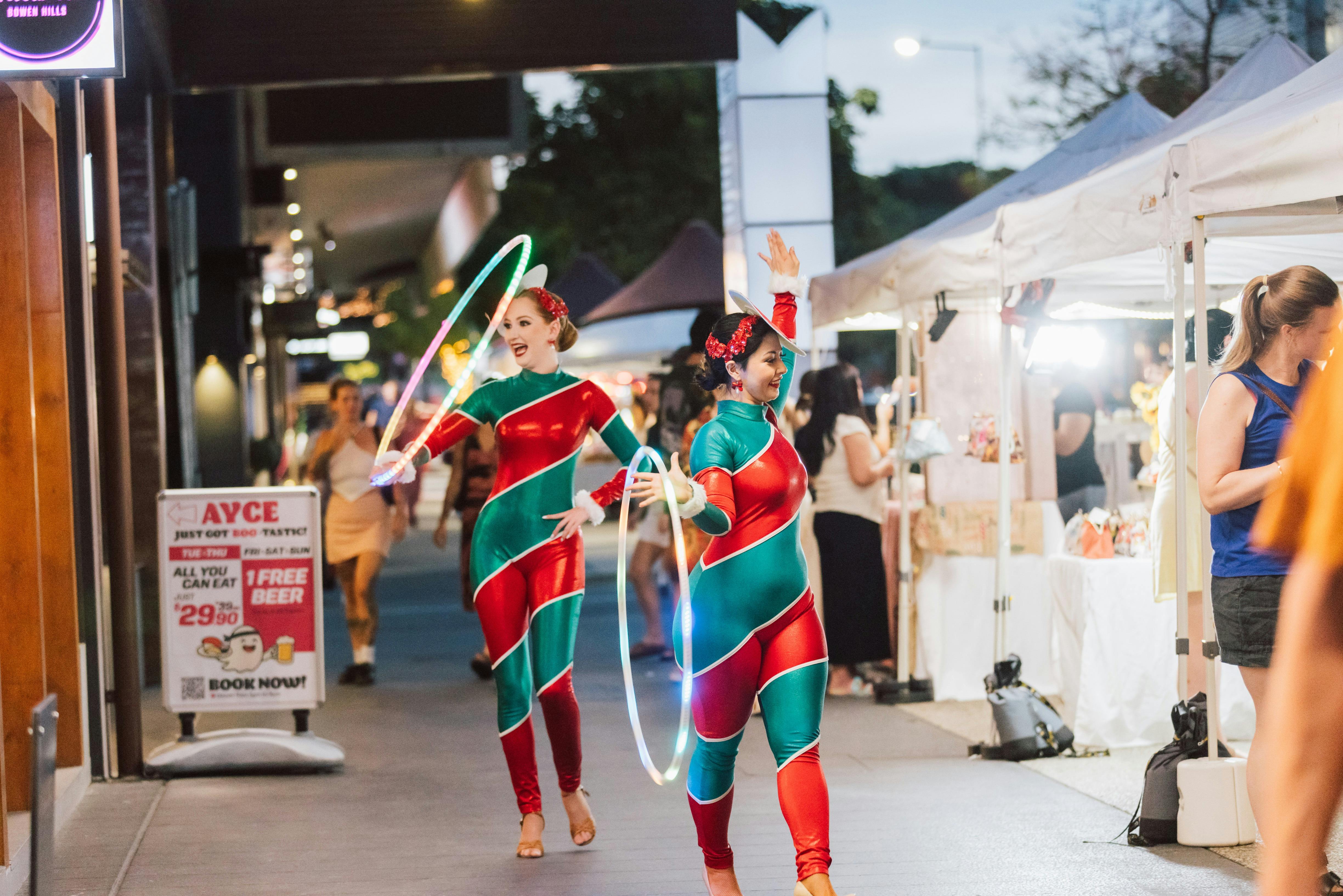 Two performers in festive red and green costumes dance with illuminated hoops at a bustling evening