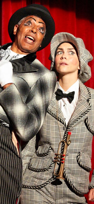 Two people in suits and hats stand in a spotlight against a red curtain, looking into the distance