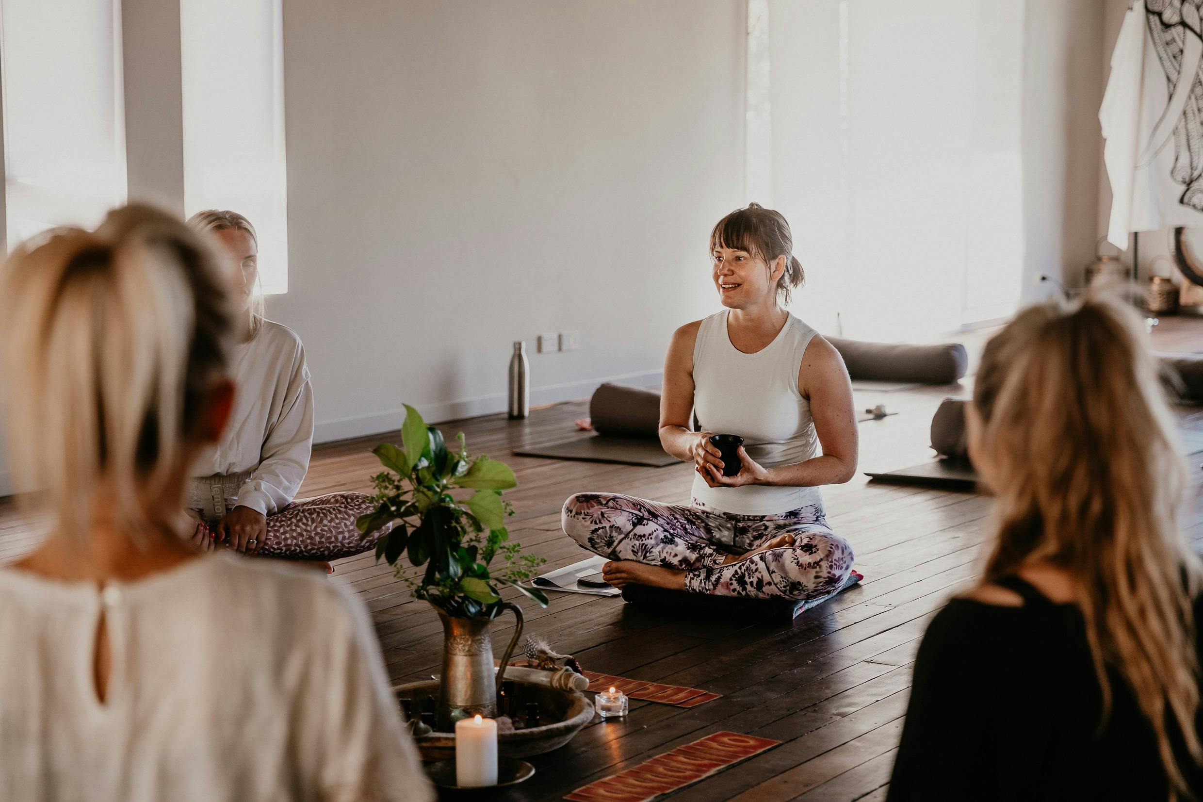 A woman in a light aqua singlet and purple patterned leggings sits on the floor in a women’s circle
