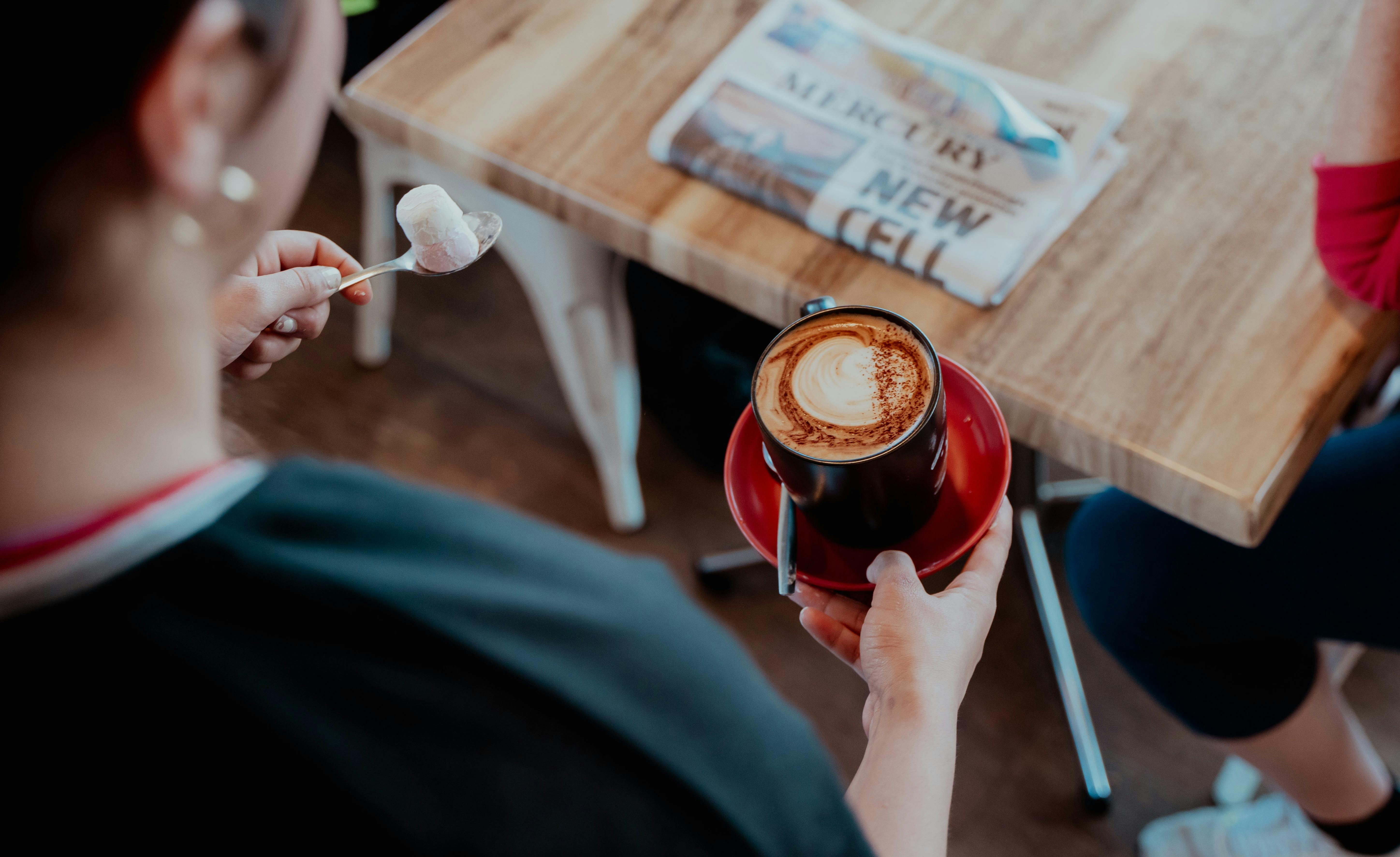 A barista holding a freshly poured coffee near a cafe table with a local paper on it