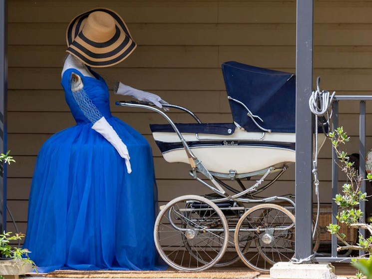a scarecrow dressed in a blue ballgown and white evening gloves wearing a floppy hat, pushing a pram