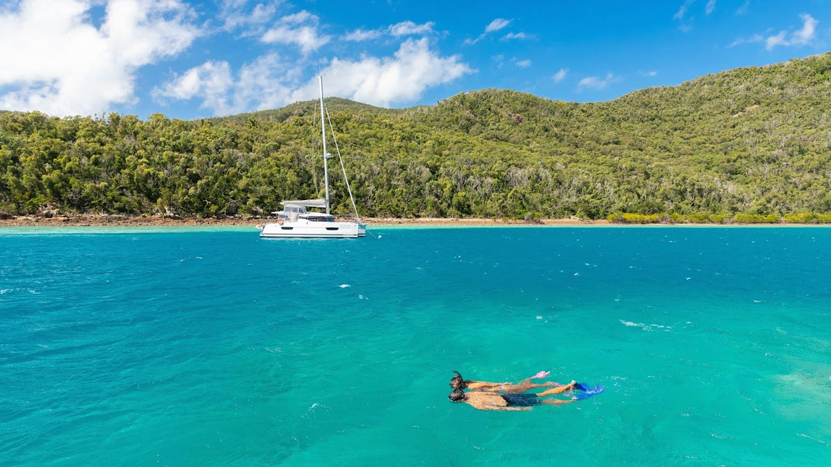 Two people snorkelling above coral reef with beach and island with trees and a sailing boat nearby