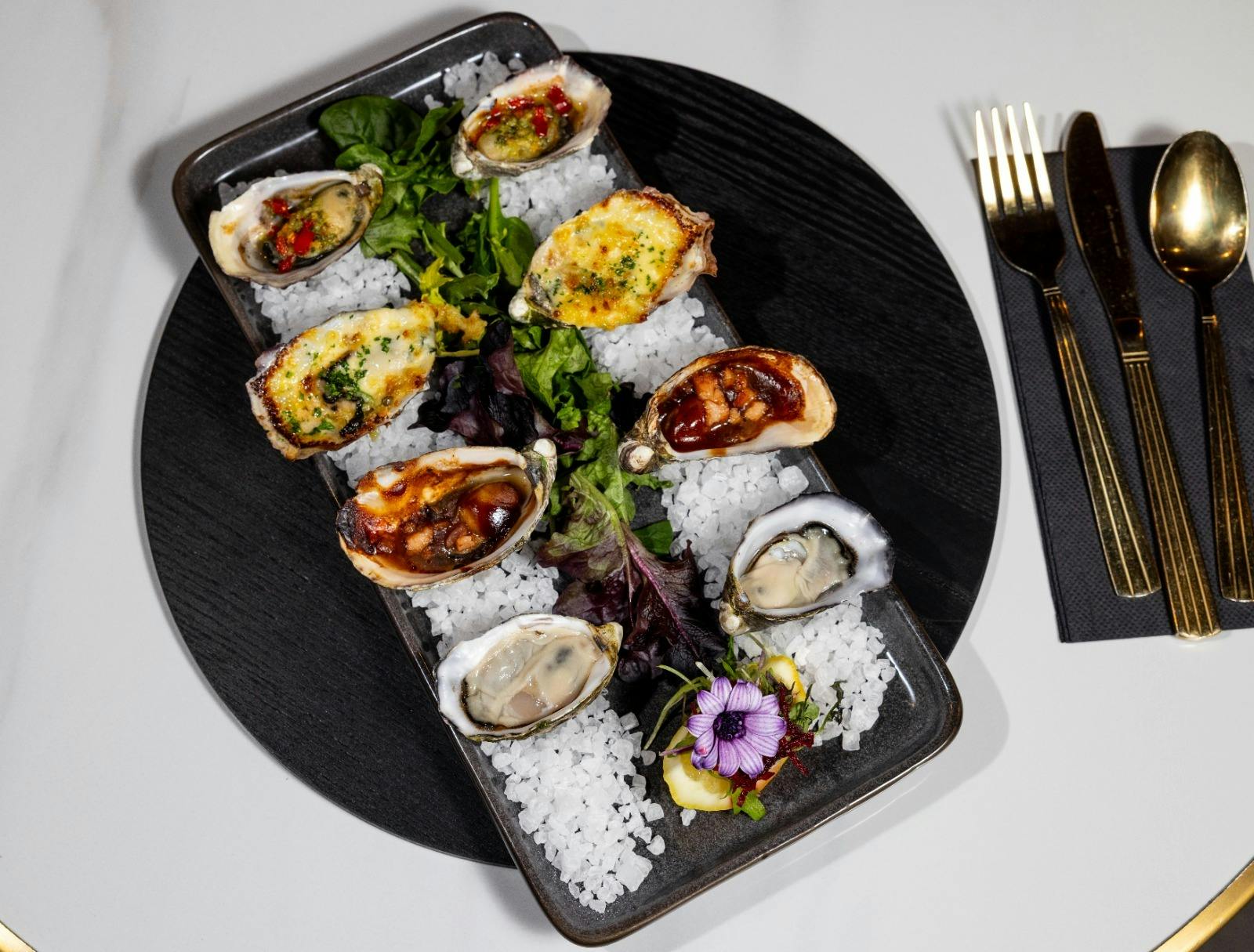Assorted fresh and baked oysters beautifully presented on a bed of rock salt with fresh greens and e