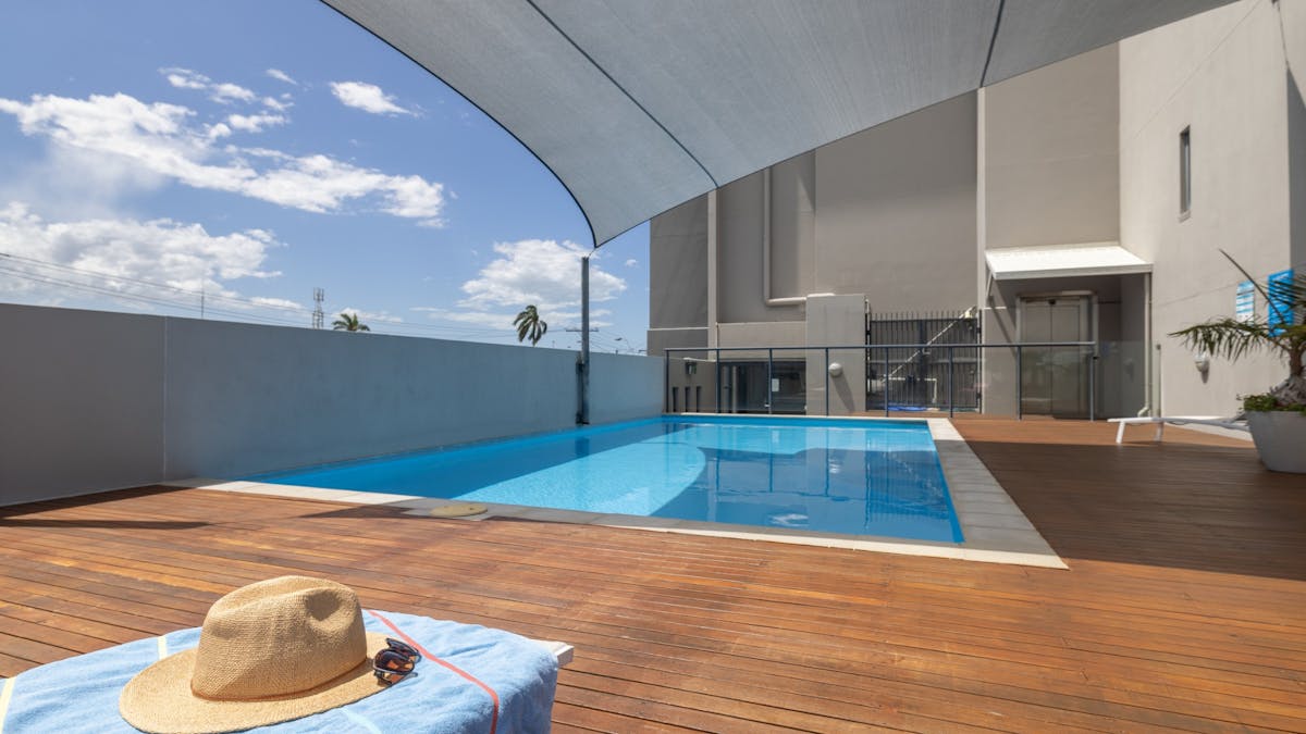 Swimming pool with wooden deck surrounding two sides. Towel, hat and sunglasses on a pool lounge.