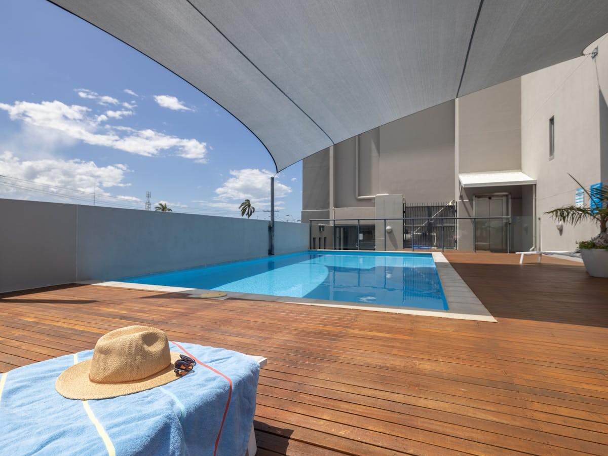 Swimming pool with wooden deck surrounding two sides. Towel, hat and sunglasses on a pool lounge.