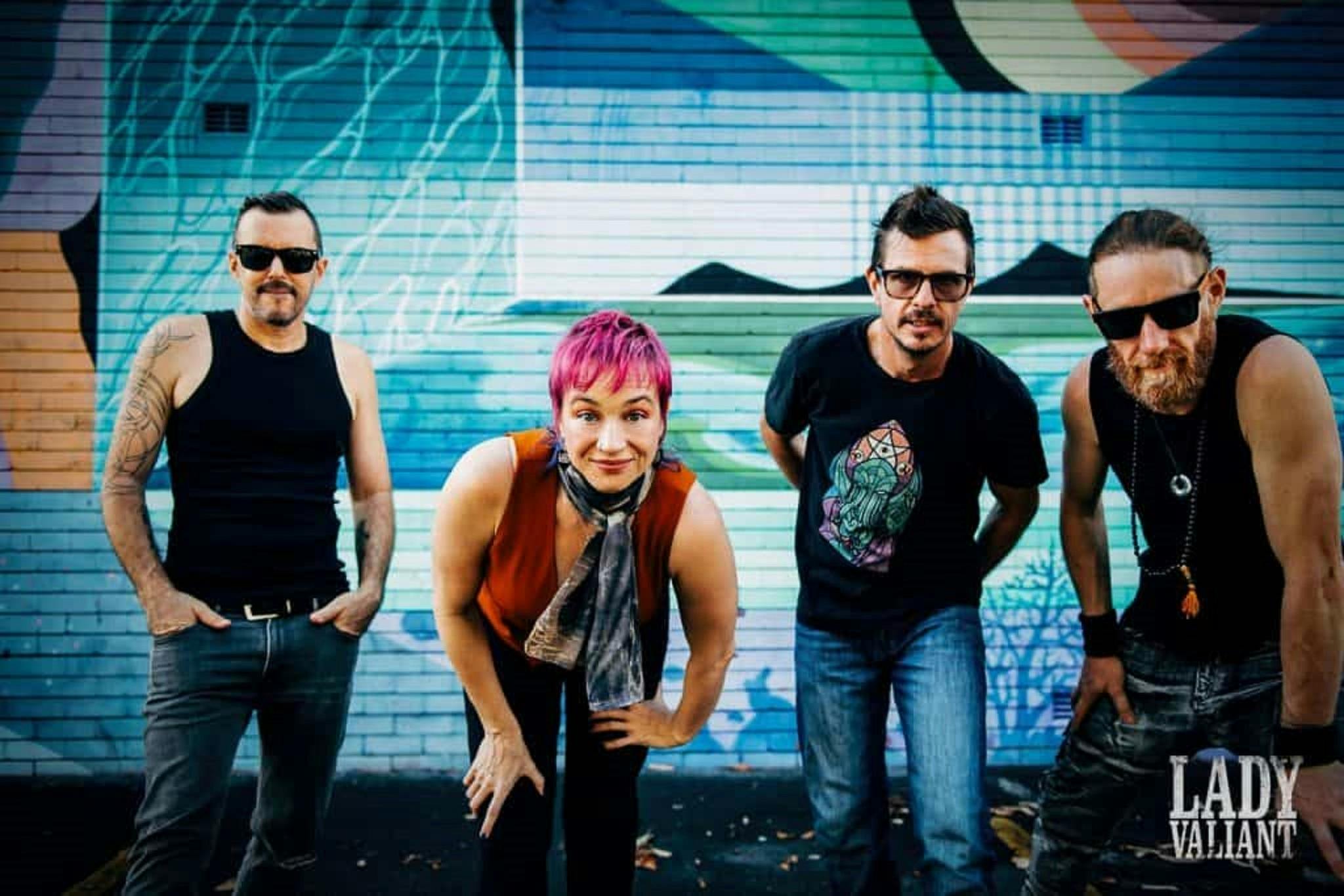 four musicians from the band Lady Valiant leaning into the camera, blue mural in backgroundd