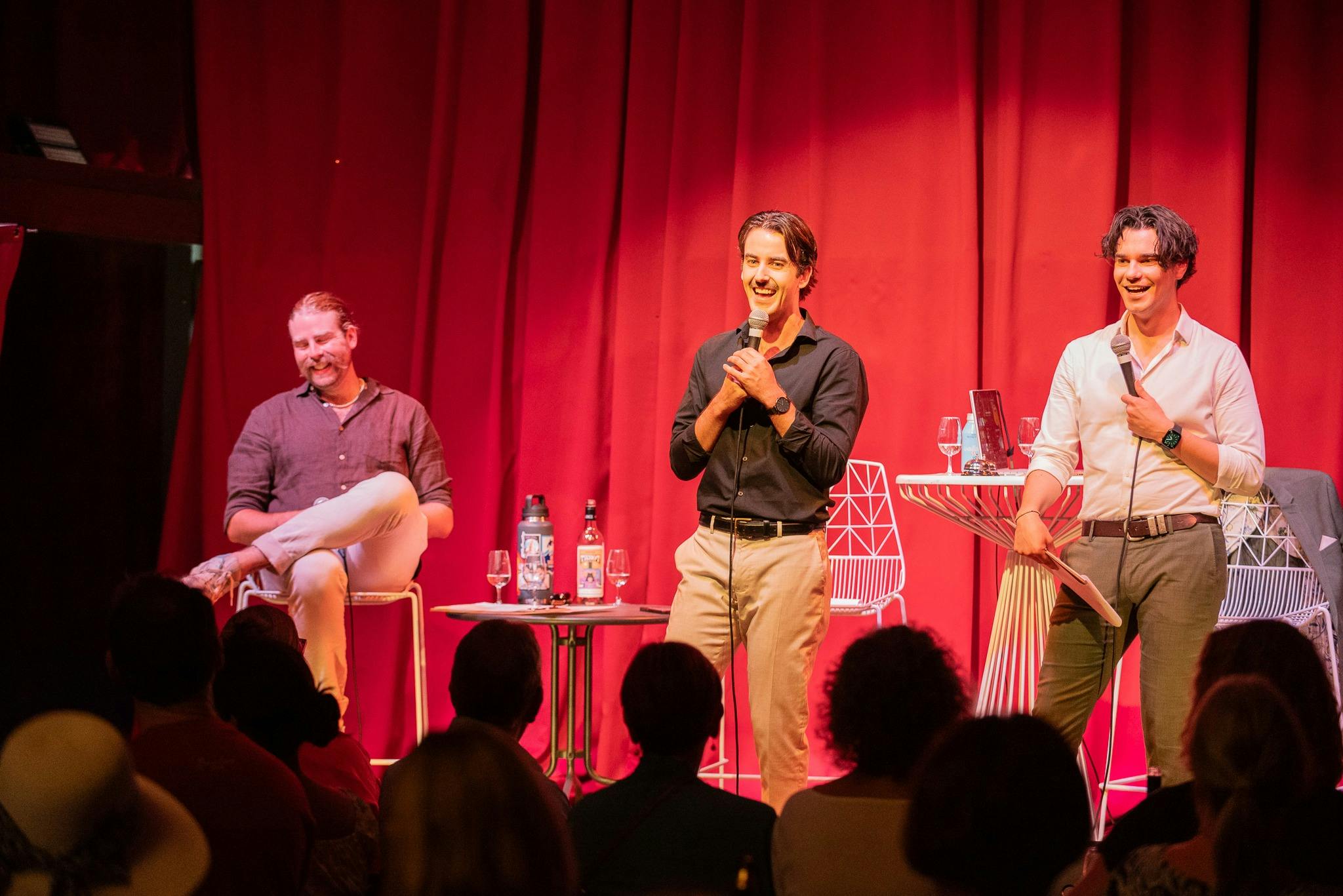Three men on stage in front of red curtain, one seated and two standing