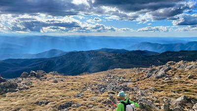 A hiker looking out into the mountains with the sun shining through the clouds.