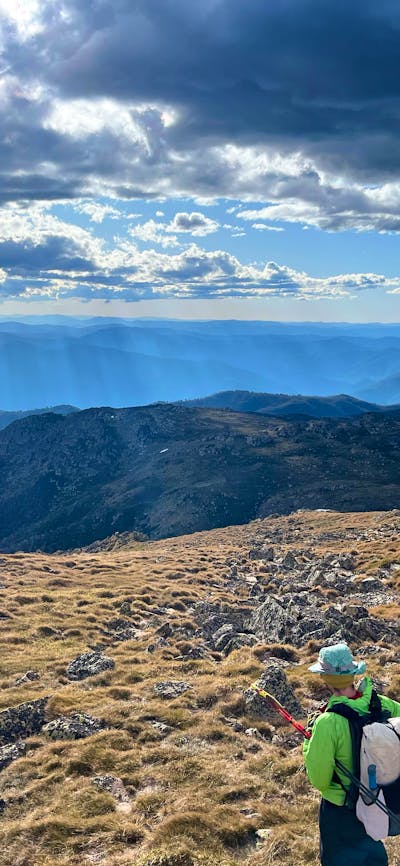 A hiker looking out into the mountains with the sun shining through the clouds.