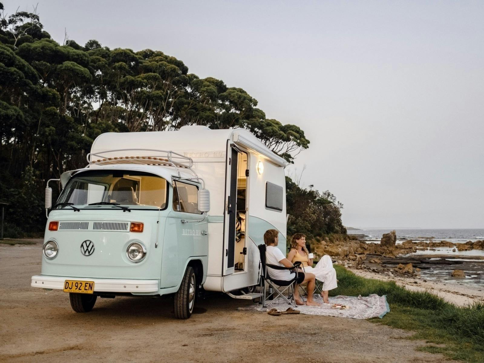 A couple sits in front of their retro campervan, beachside