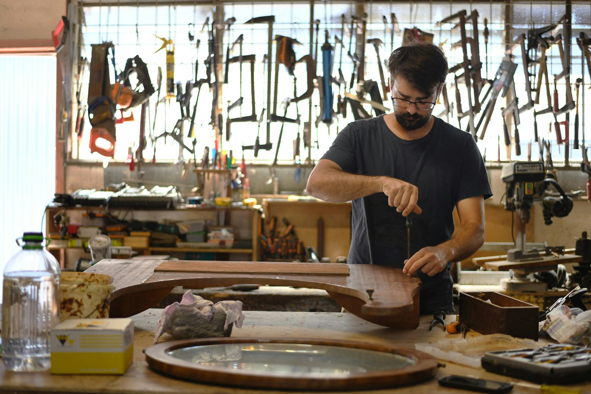 Craftsperson working on wooden furniture with a hand tool in a workshop filled with tools.