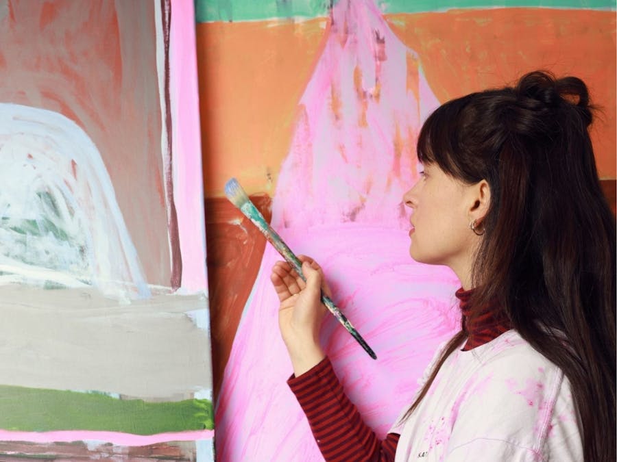 Artist holds paint brush whilst standing with several artworks behind her