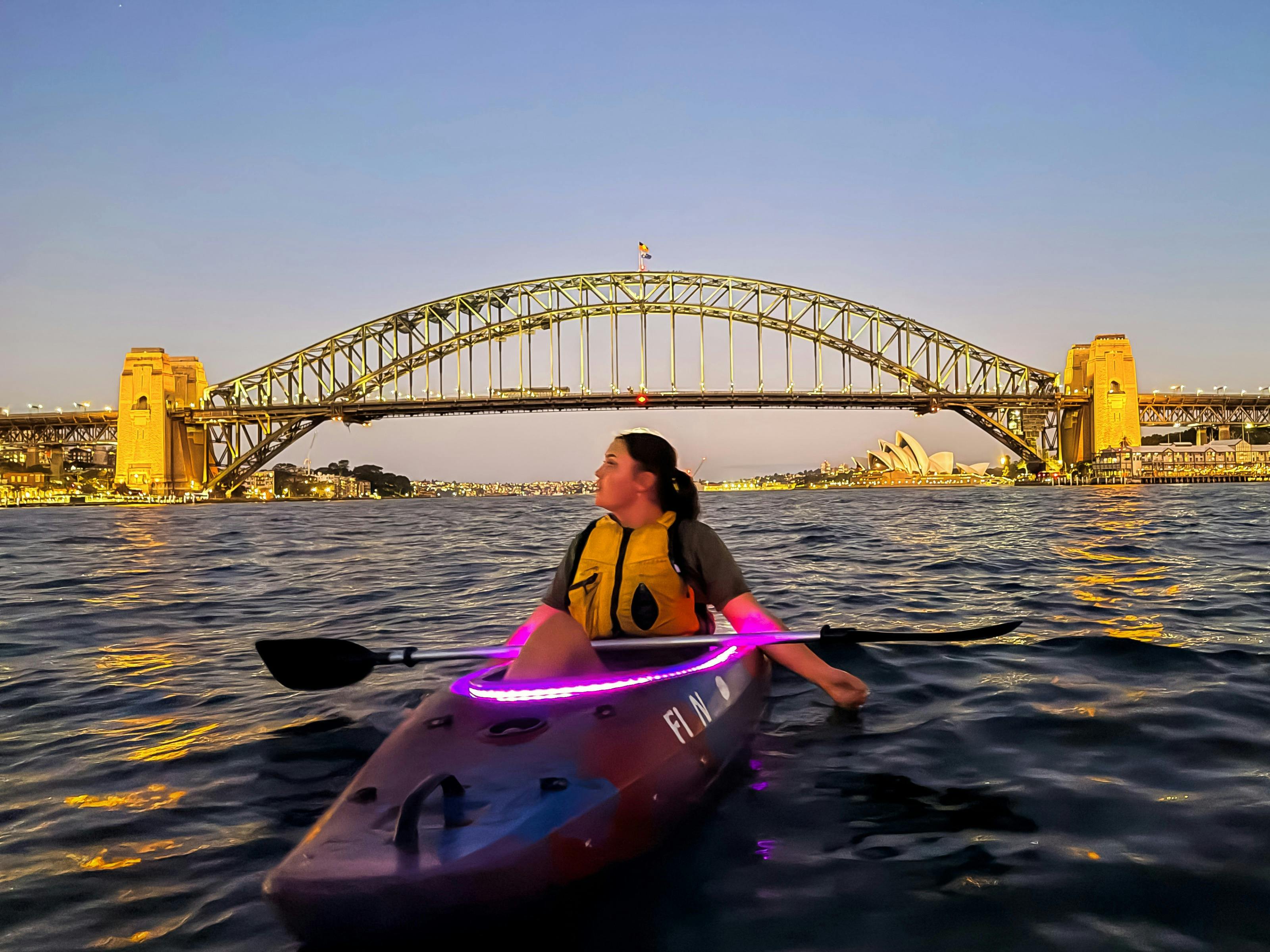 Girl watches Opera House and Sydney Harbour Bridge glow at sunset from her pink-led-lit kayak.