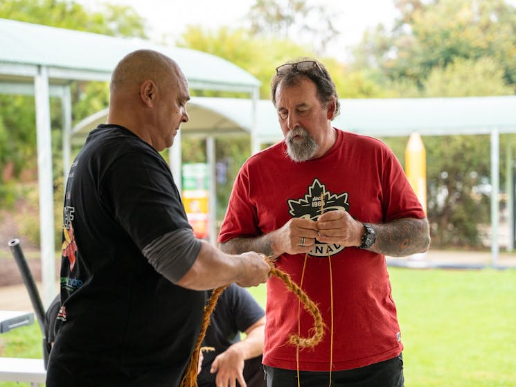 An Aboriginal man in a black tshirt showing another man how to make rope using traditional methods.