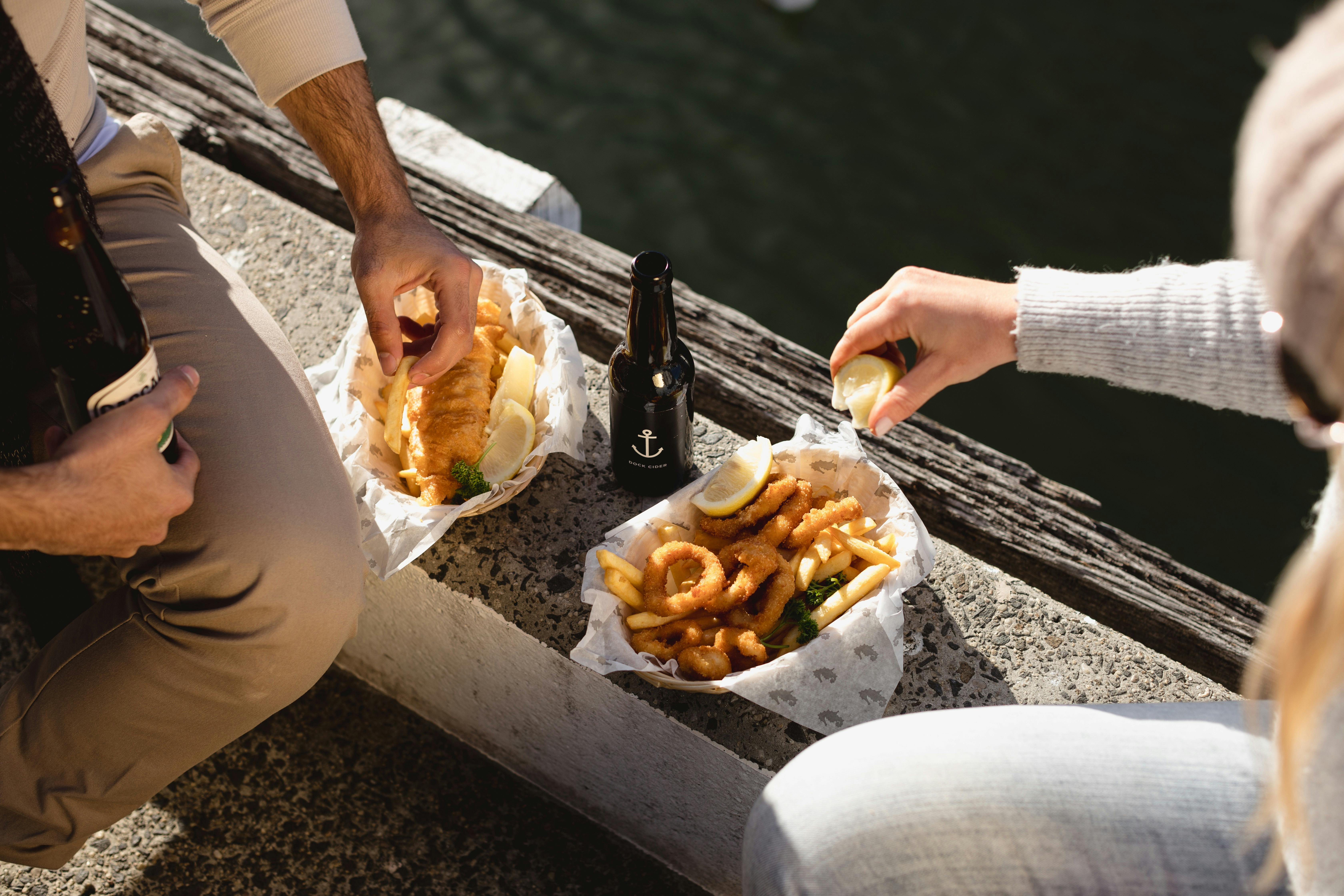 Two people enjoying fish and chips on the side of the dock outside Mures Lower Deck.