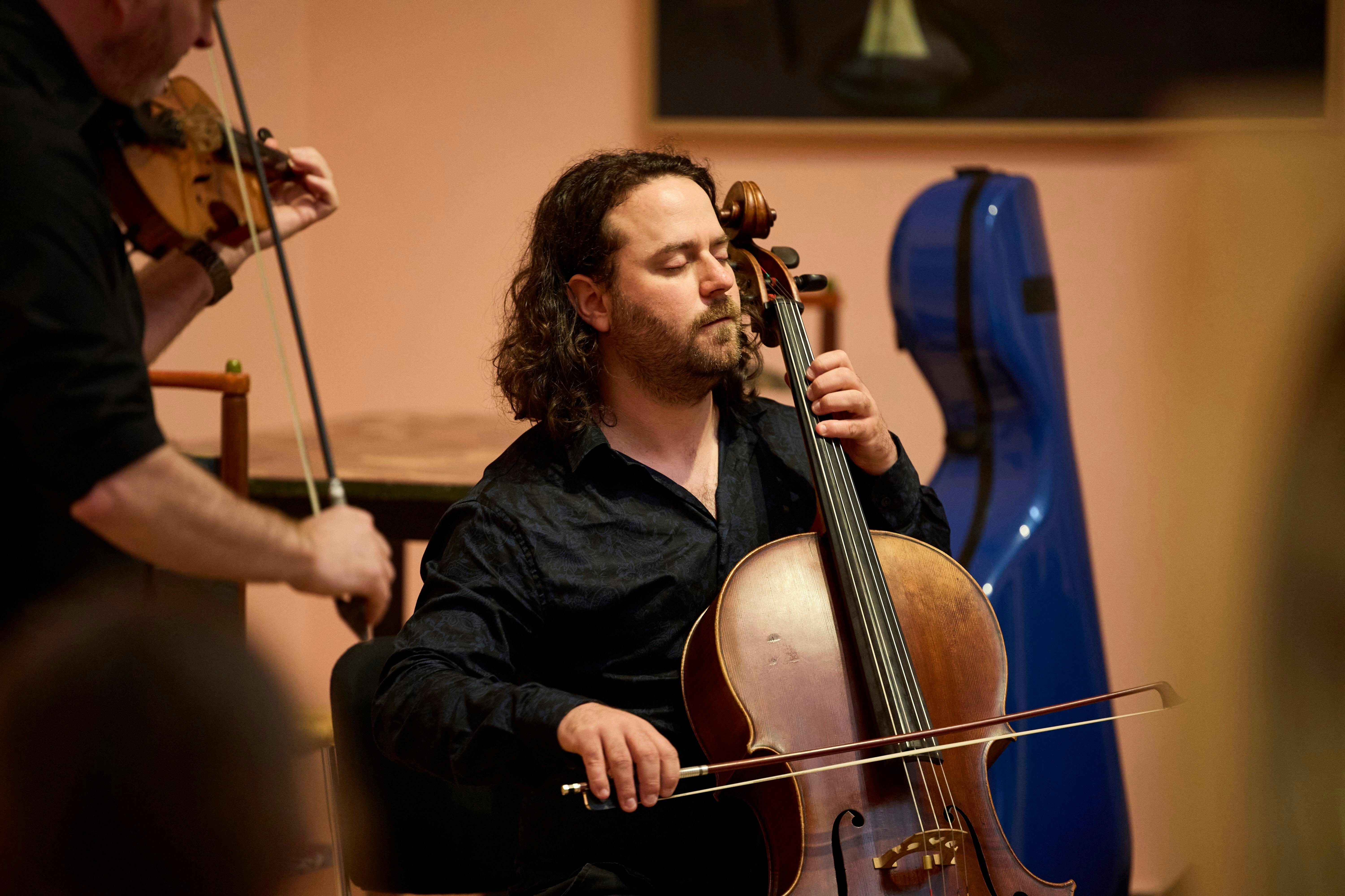 A violinist and cellist perform in an art gallery. The cellist is seated with their eyes closed.