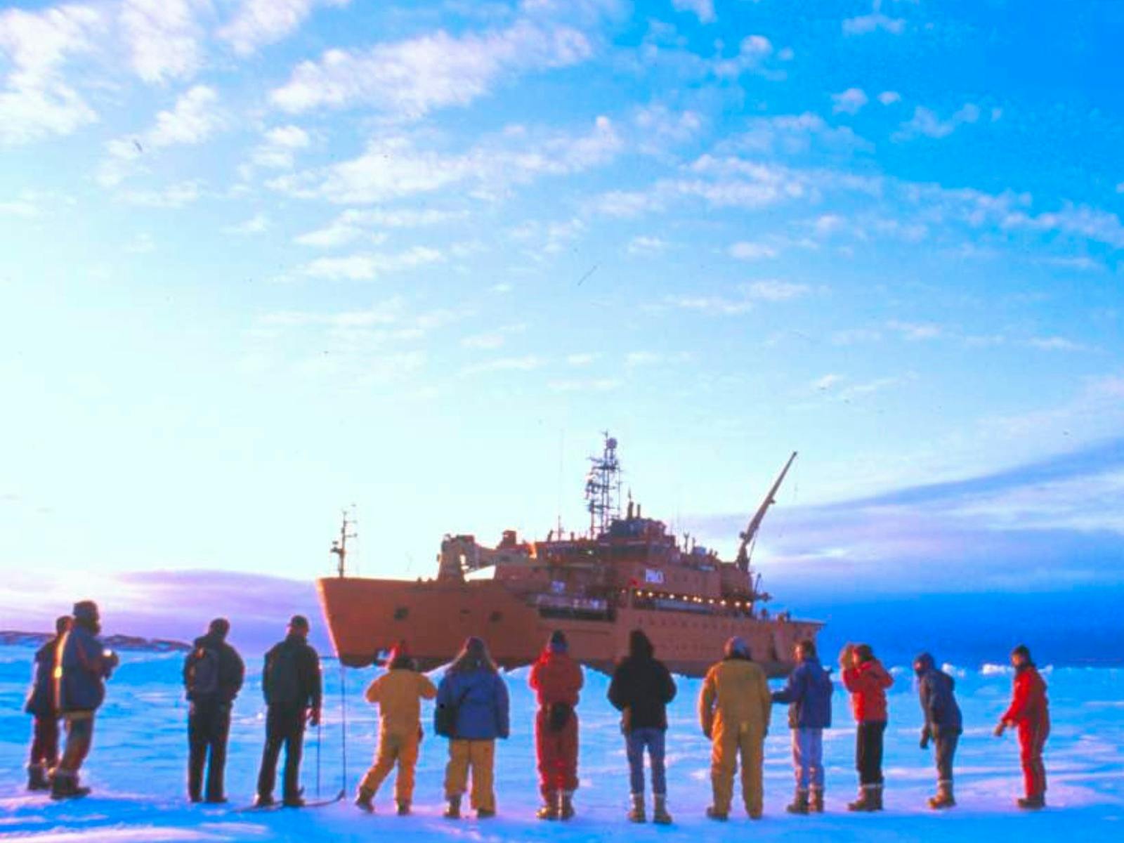 People in colorful winter gear stand on a vast icy plain, facing a large orange icebreaker ship.