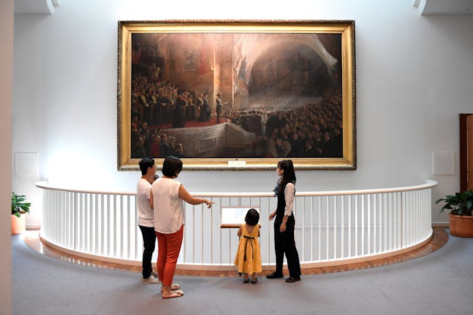 Visitors stand on an upper level of Parliament House, viewing a large framed historical painting.