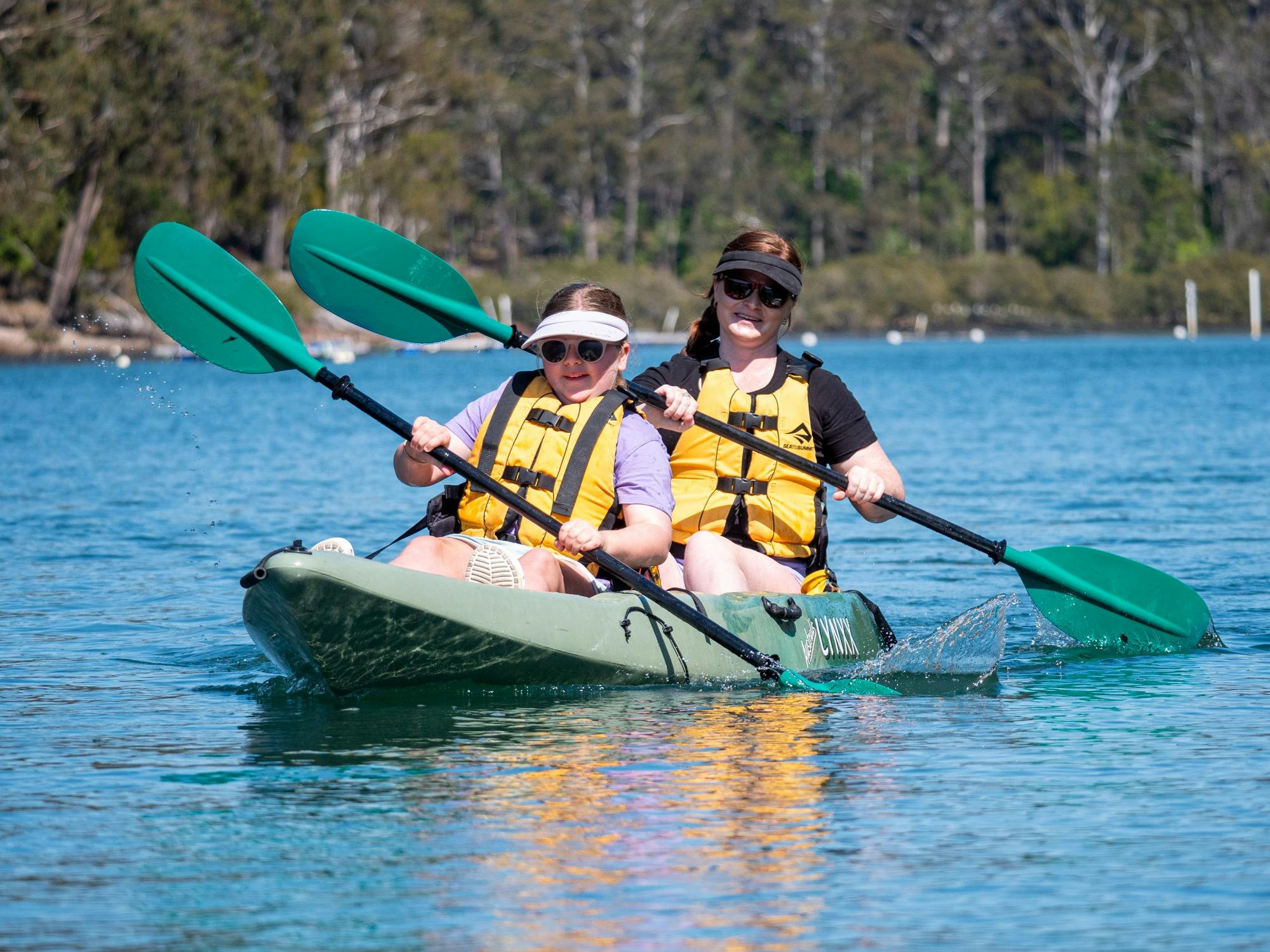 A mother and daughter in a green kayak, wearing hats and PFDs, smiling as they paddle in sync