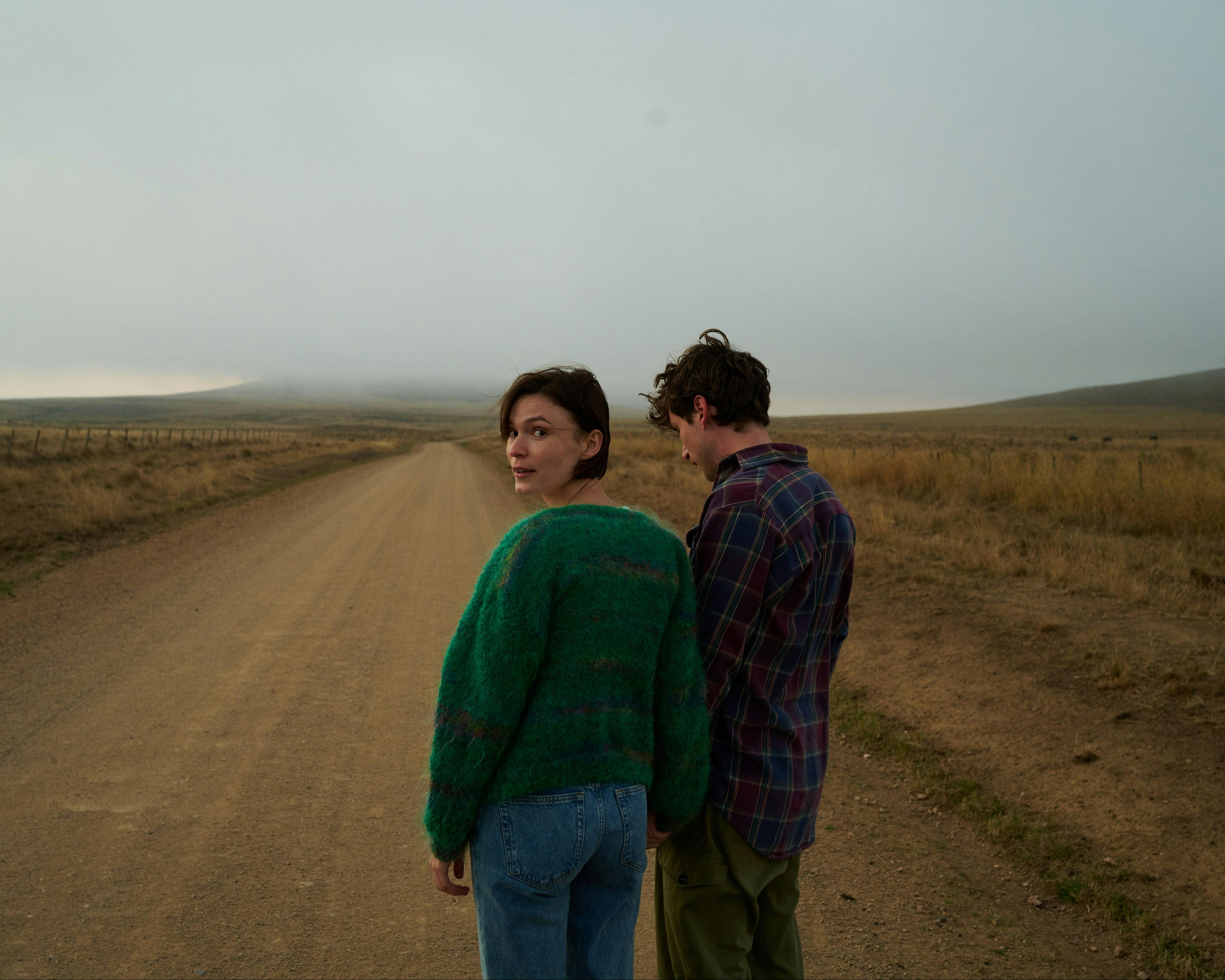 A woman in a green jumper and a man in a plaid shirt hold hands on a country road