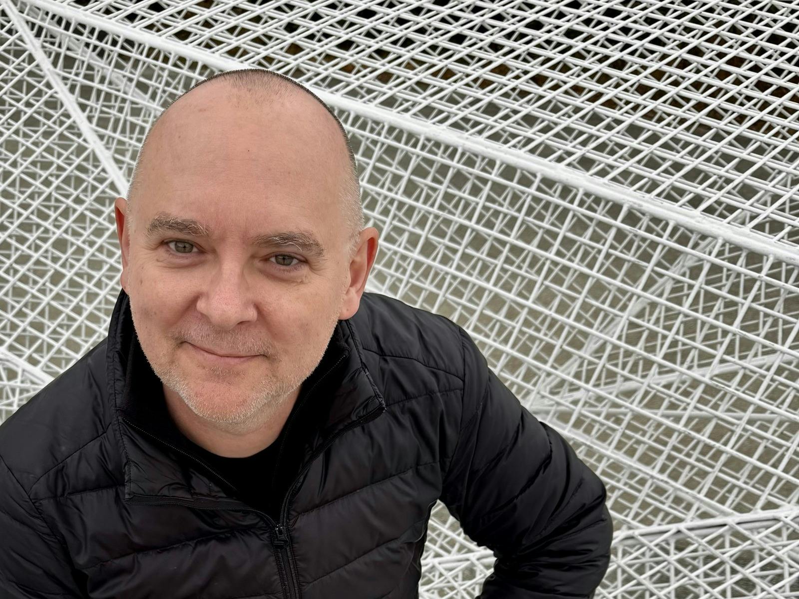 Man in black jacket smiles against white geometric mesh background.