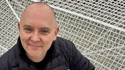 Man in black jacket smiles against white geometric mesh background.
