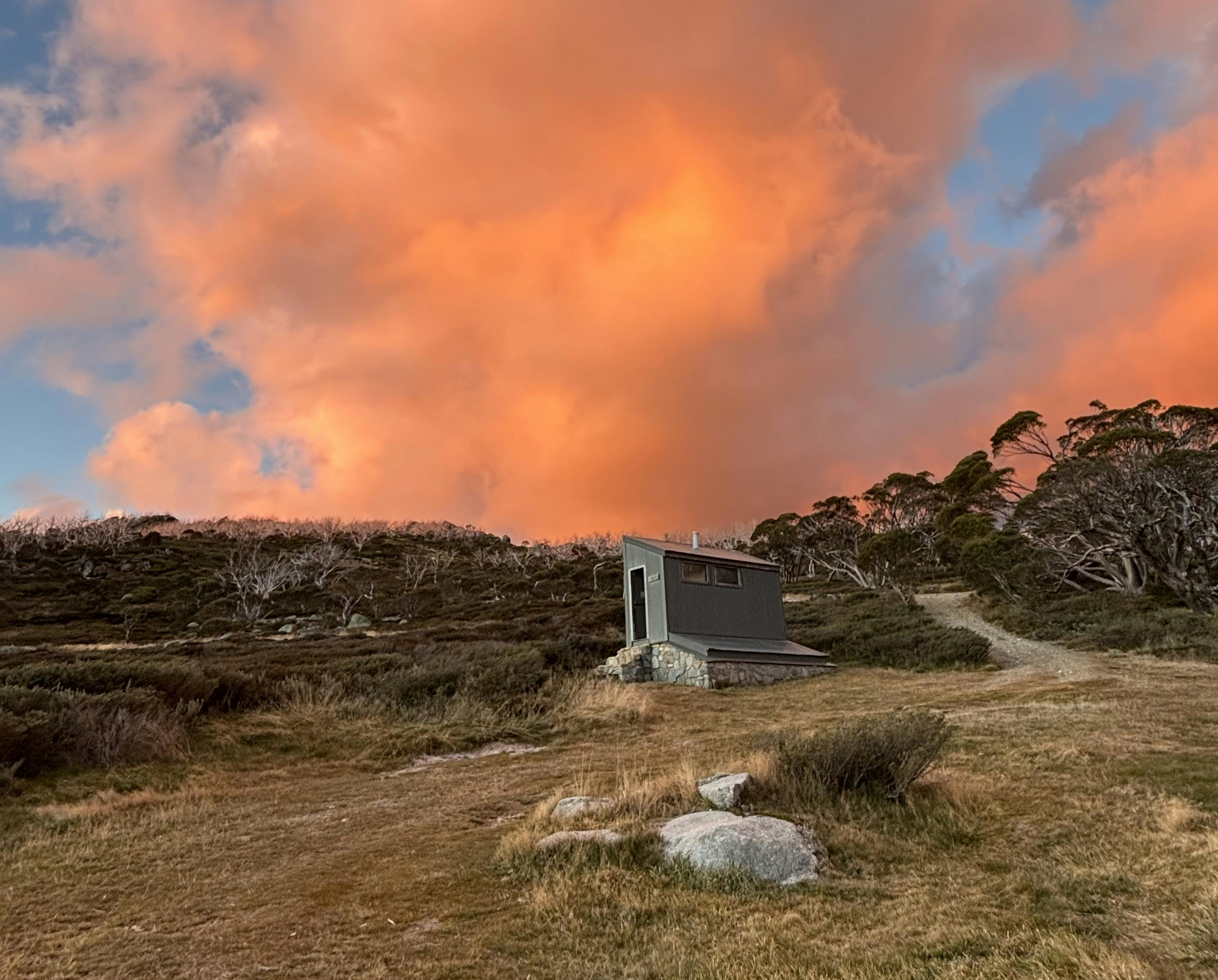 A small green hut bathed in a golden sunset light. Orange clouds and blue sky above.