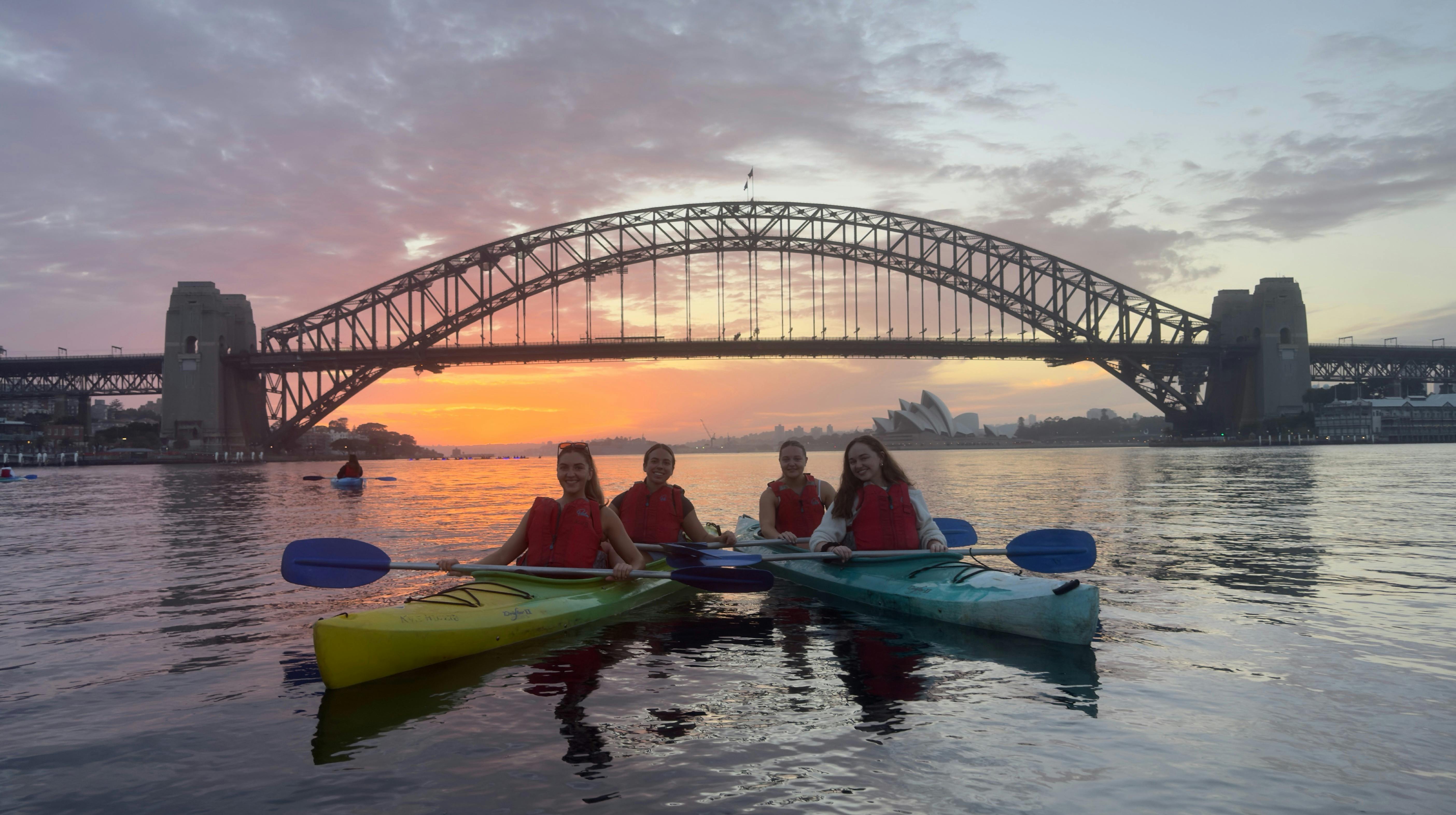 一群皮划艇愛好者在雪梨港的日出中劃行，背後是海港大橋和城市天際線。