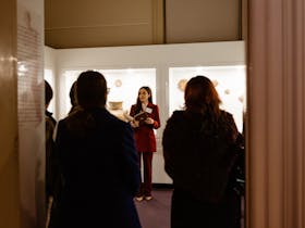 Woman in a red suit with staff badge stands in an exhibition speaking to visitors seen from behind