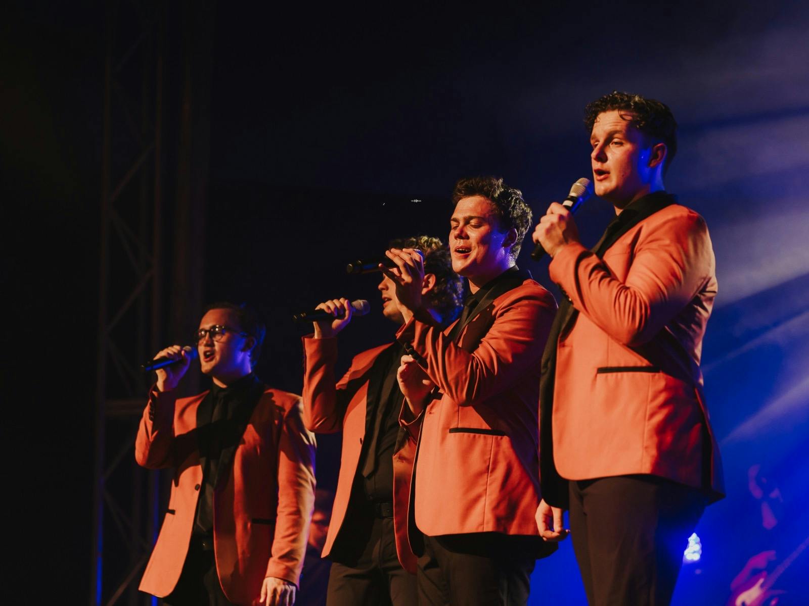 Close-up of the band singing on stage, dressed in orange jackets under dramatic stage lighting.