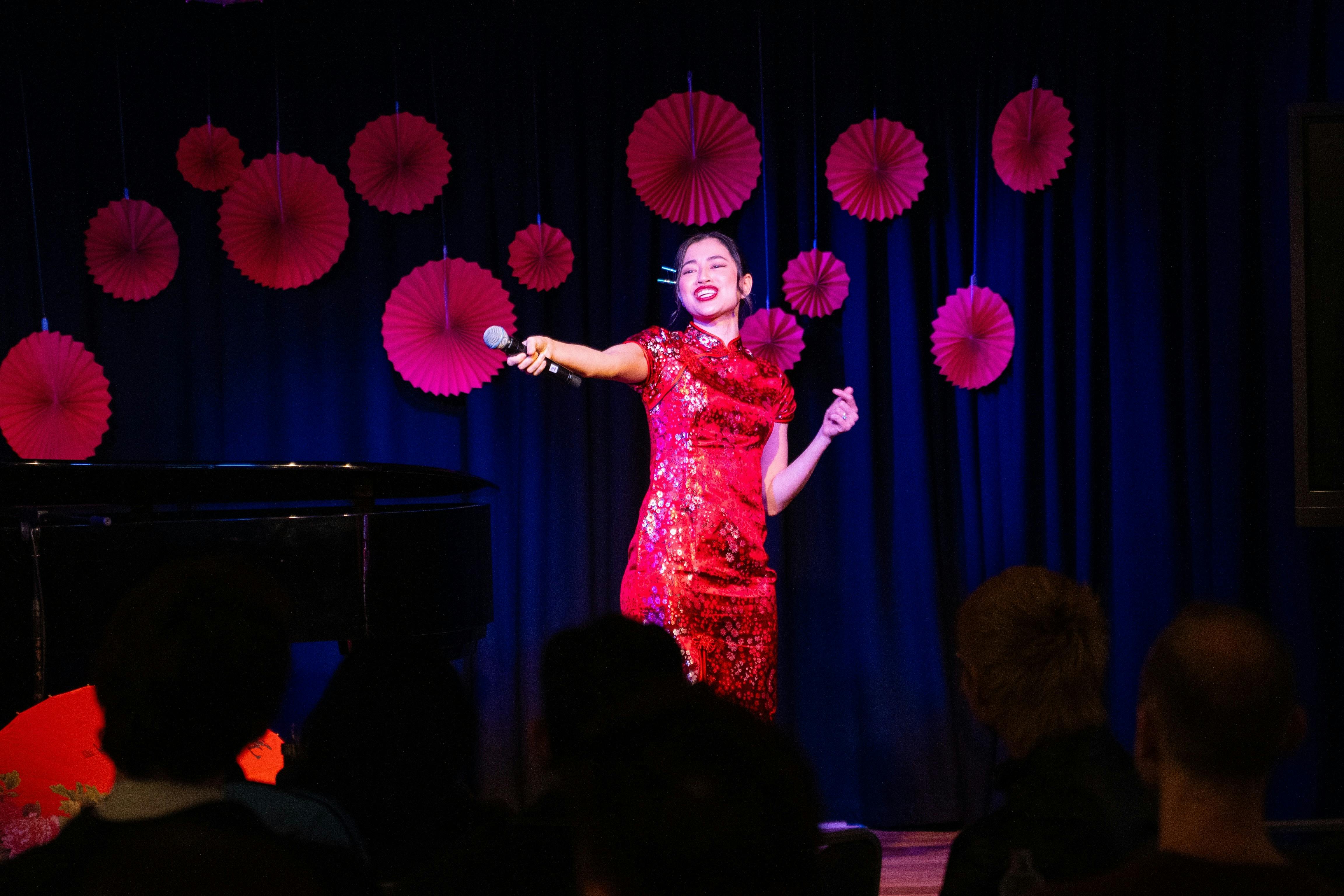 A woman in a red dress stands confidently on stage, engaging the audience with her presence.