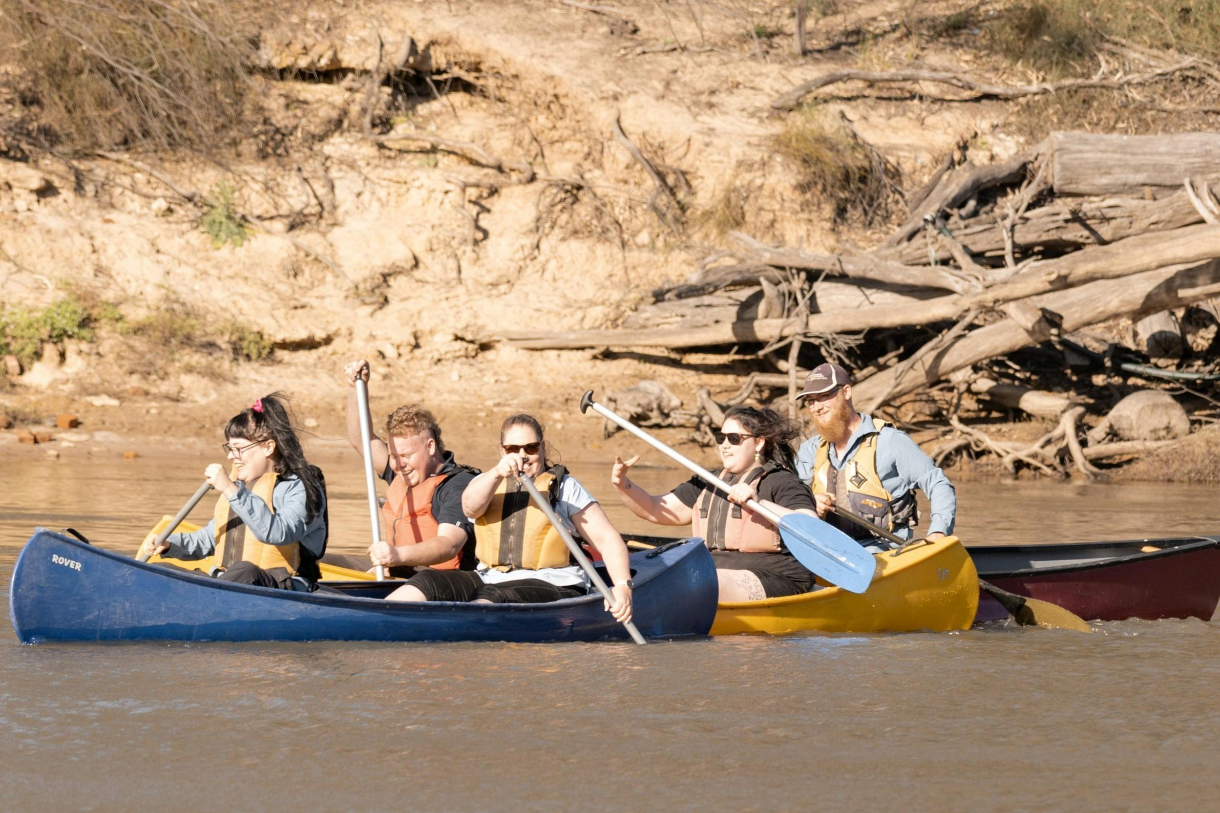 Five people wearing life jackets paddle three canoes past a sandy, tree-lined riverbank.