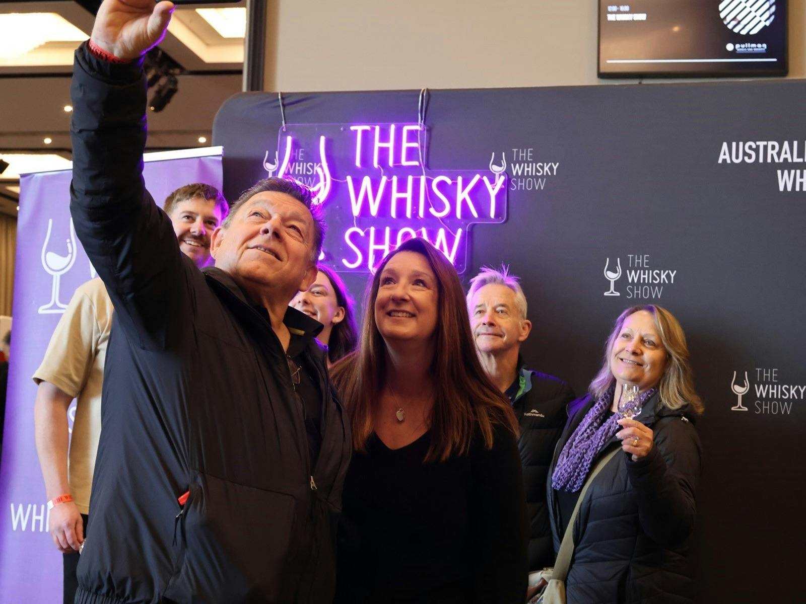 A group of friends gather in front of a Whisky Show banner and Neon Sign. A group of friends gather in front of a Whisky Show banner and Neon Sign.