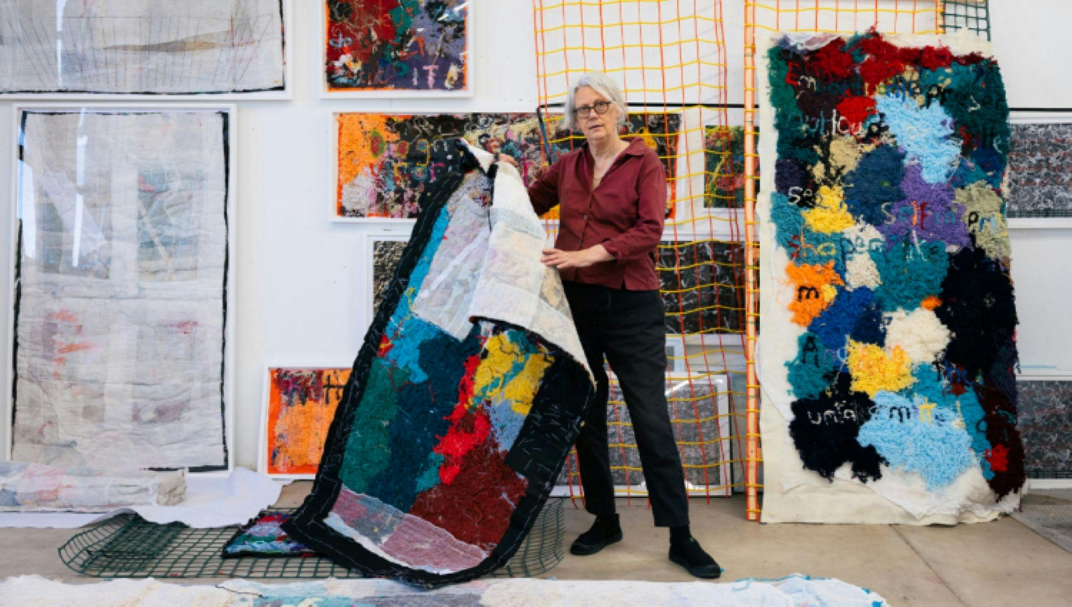 Elizabeth casually holding up a textile work, wearing a red shirt and black pants